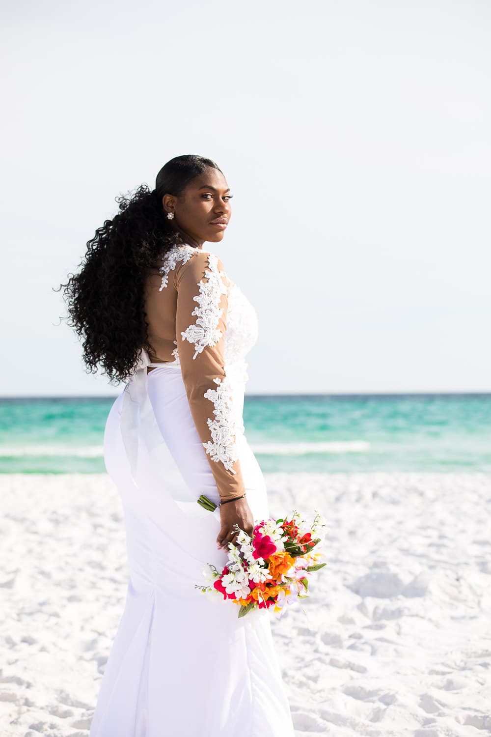 A bride standing on the beach facing the ocean and looking back at the camera.